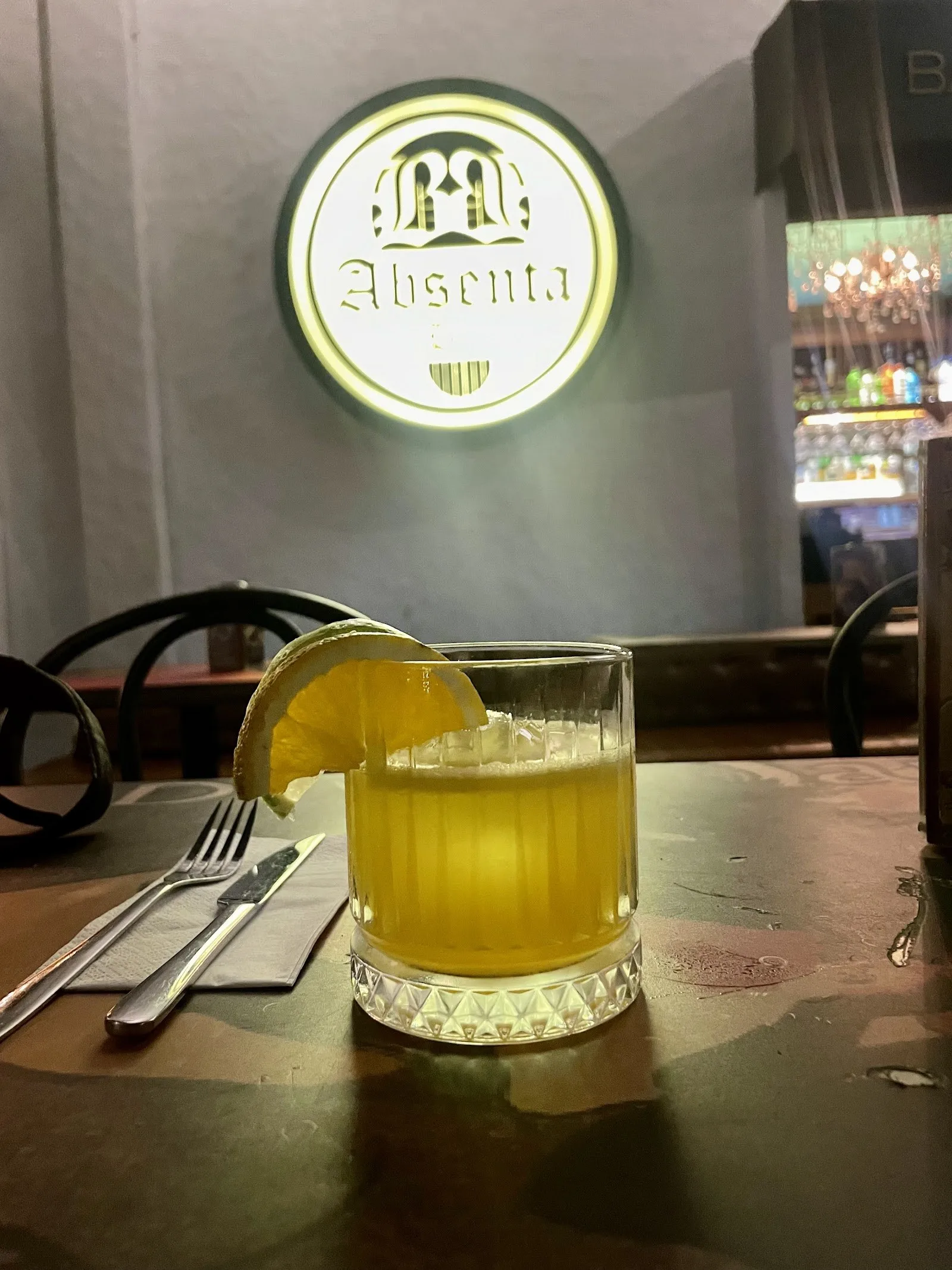 Cóctel servido en una mesa de Absenta con el letrero luminoso del bar al fondo