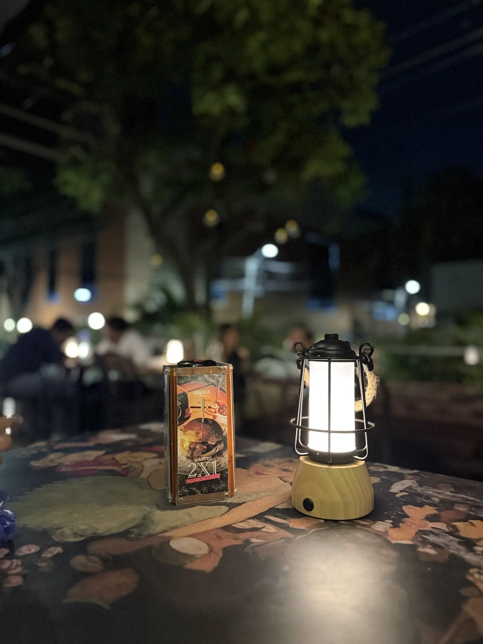 Mesa de afuera de Absenta en la noche, con farol y árboles de El Peñón