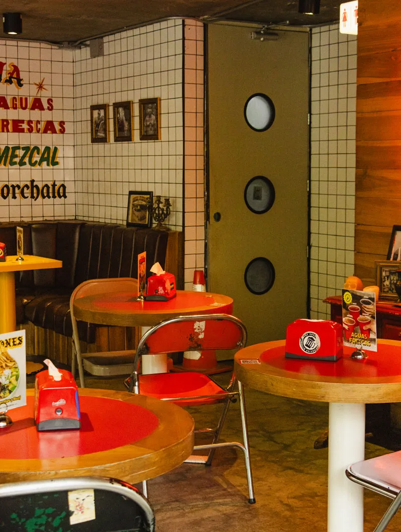 Salón de baldosa blanca con mesas rojas y letreros de aguas frescas, mezcal y horchata en Criminal Taquería