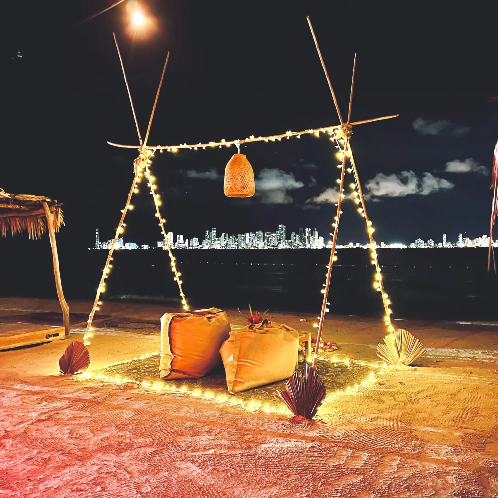 Mesa de cena en la arena de Fénix Beach con luces y cojines, con Cartagena iluminada al fondo