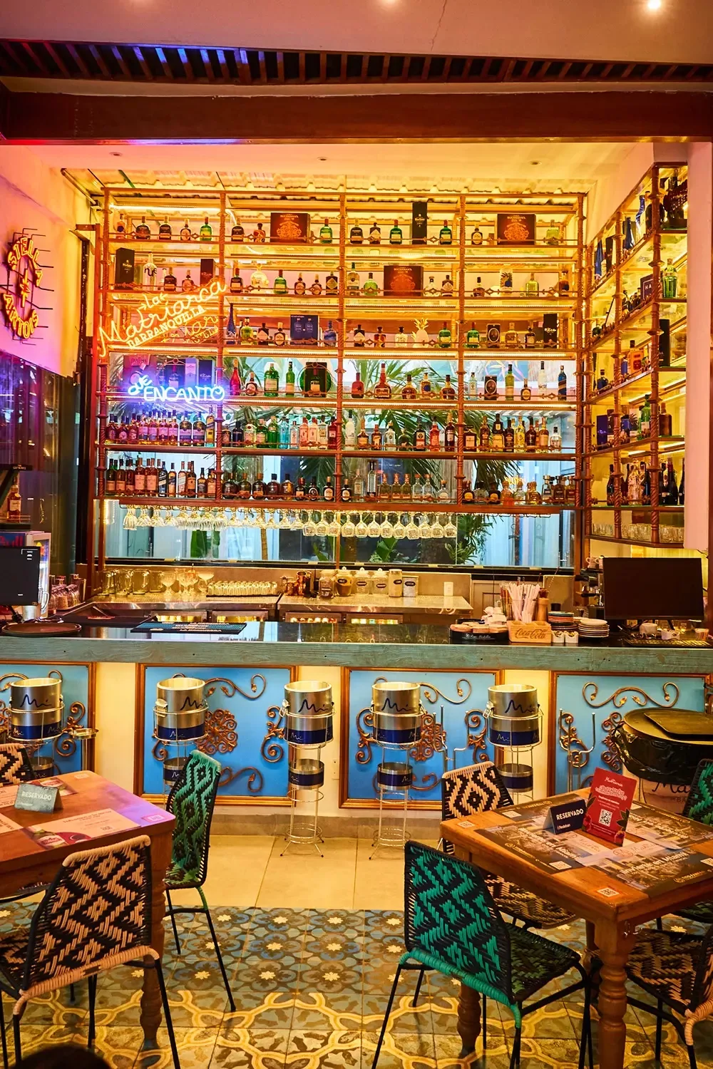 Bar iluminado de La Matriarca con estantería de botellas, baldosa de colores y sillas tejidas