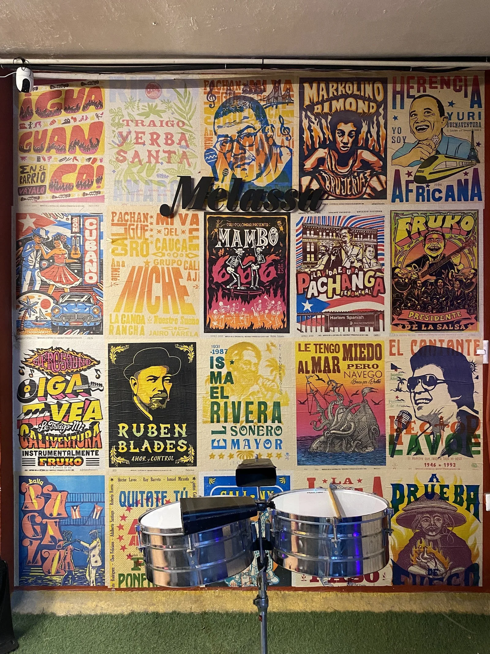 Pared de afiches de orquestas de salsa en Melassa, con timbales al frente
