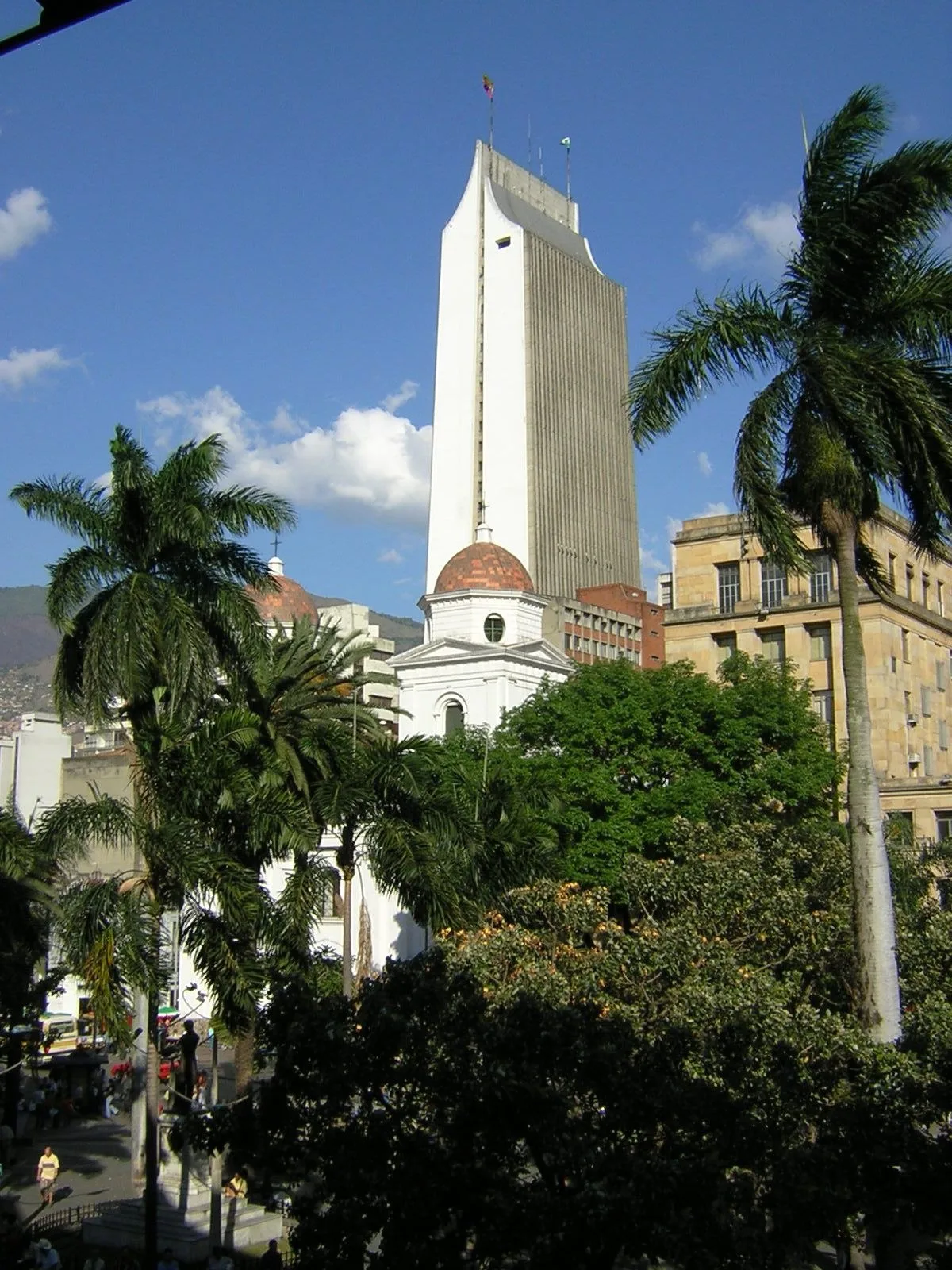 Edificio Coltejer y la fachada blanca de la Basílica de la Candelaria entre palmas, vistos desde el Parque Berrío