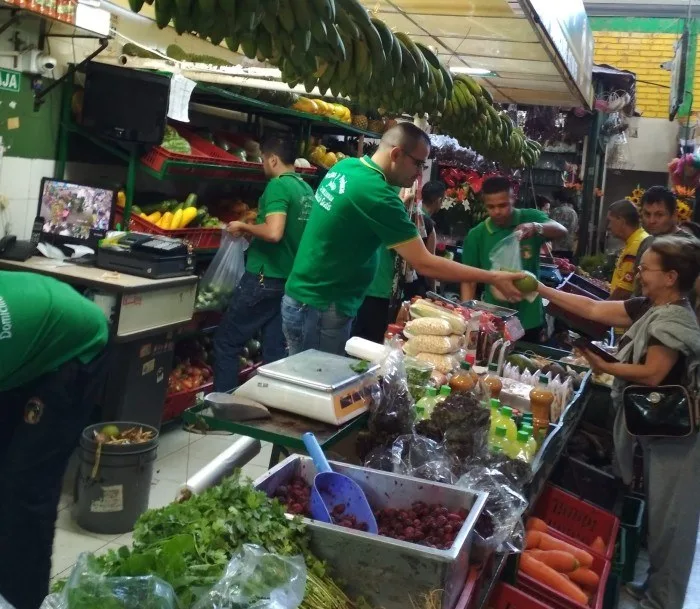 Venteros de camiseta verde atendiendo clientas en un puesto de verduras y frutas de la plaza