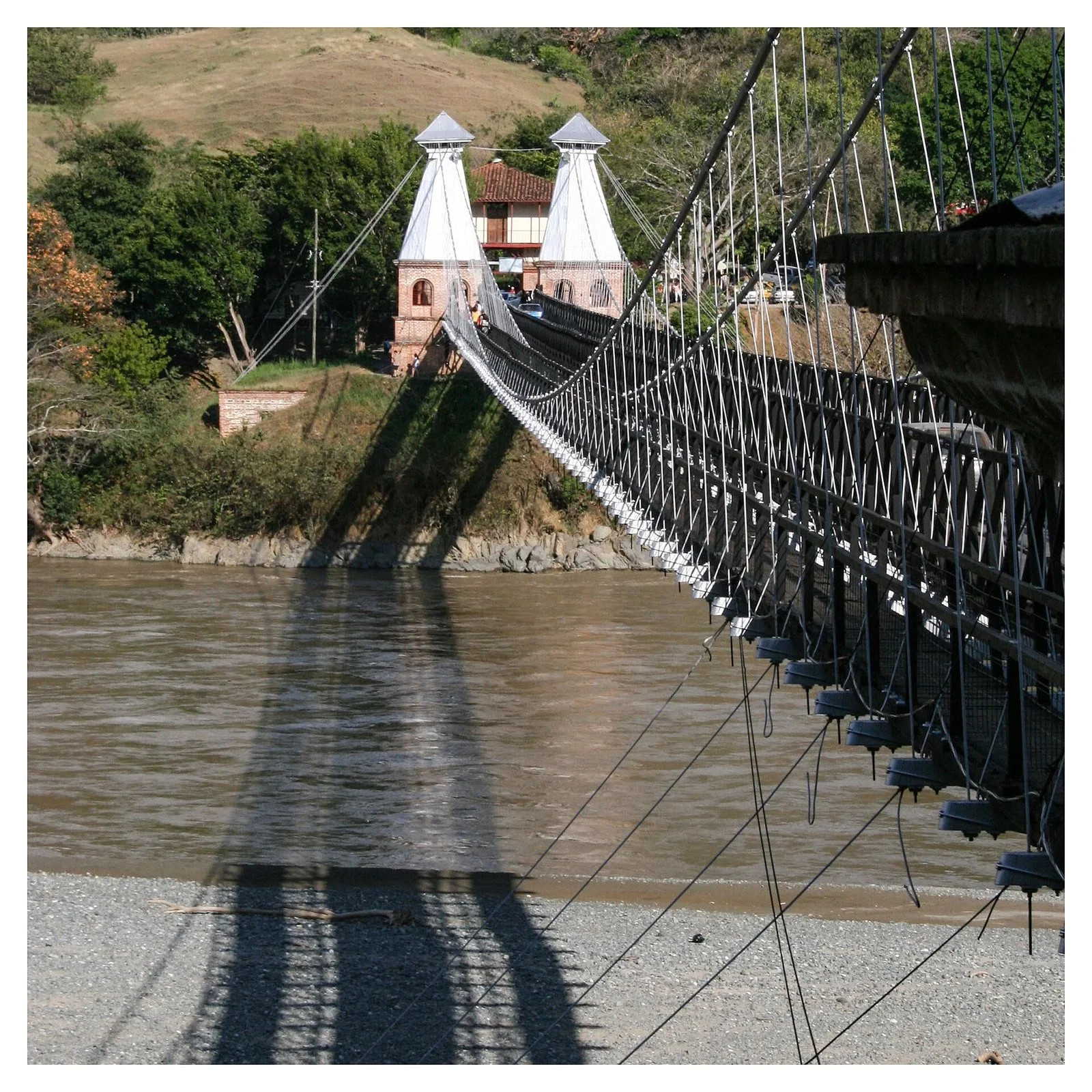 Costado del Puente de Occidente con sus cables y la sombra sobre la playa del río Cauca, y las torres blancas al fondo