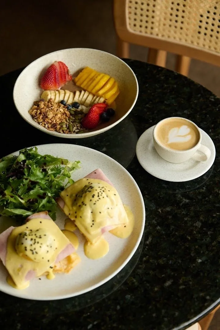 Huevos benedictinos, bowl de fruta con granola y capuchino sobre mesa de mármol